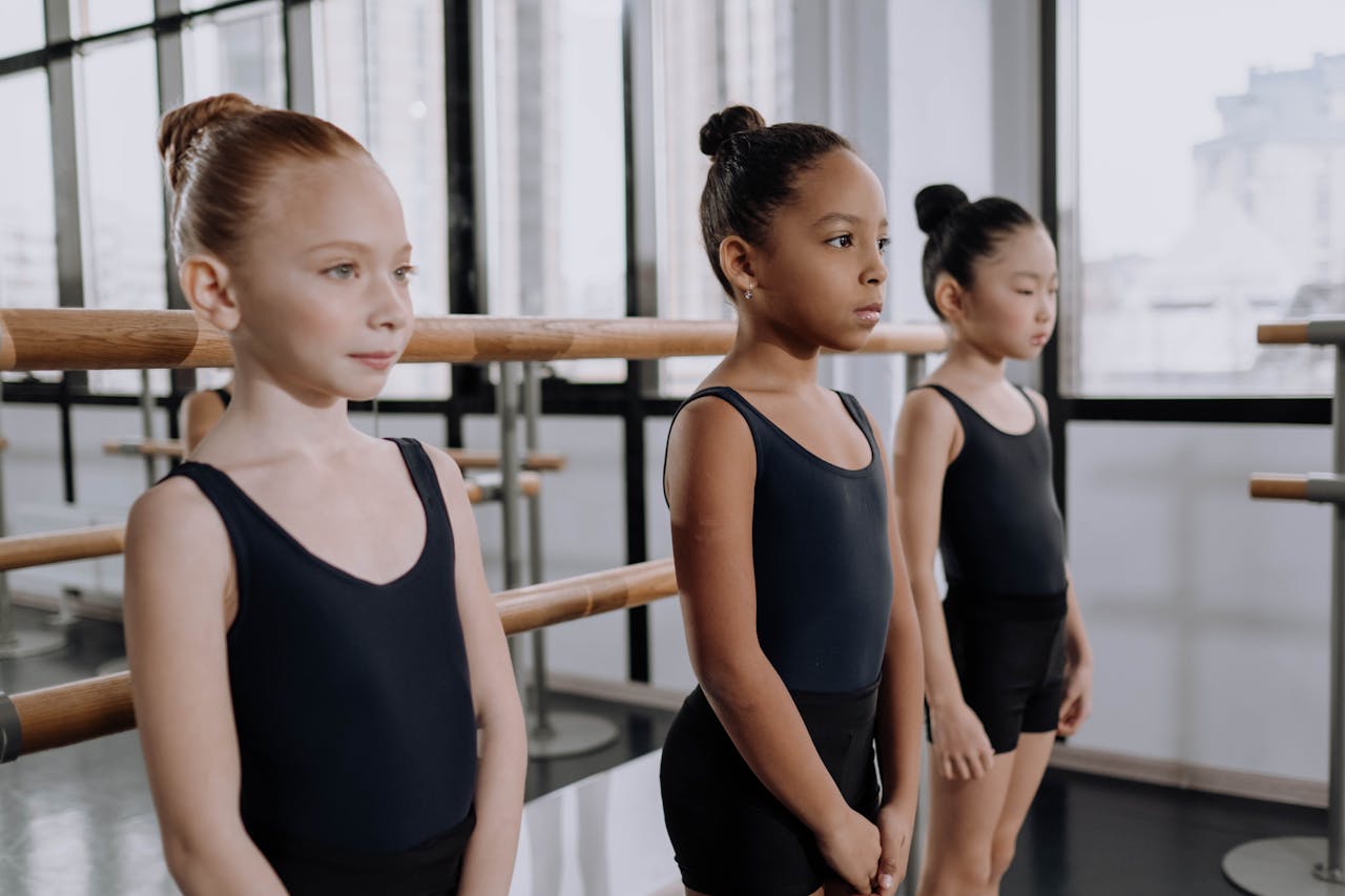 Three young girls in ballet outfits practicing in a well-lit dance studio with determination.